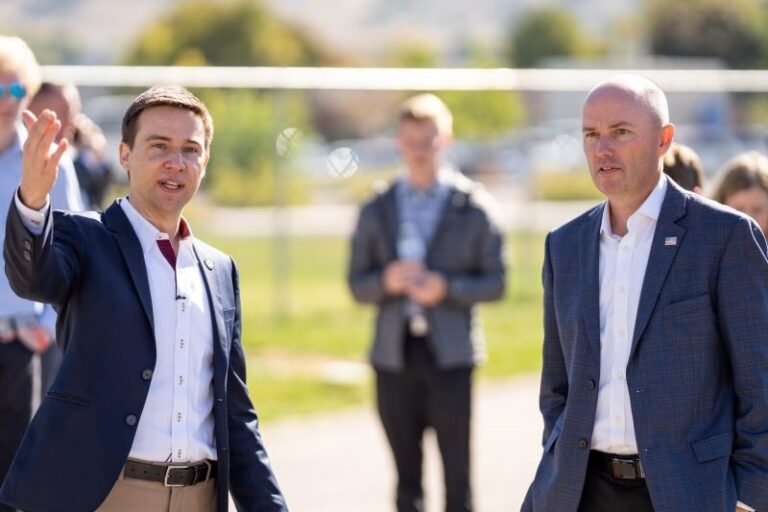 Two men in suits speaking outdoors, one gesturing, at an outdoor event