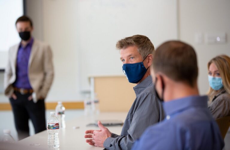 Masked man in a meeting speaking to colleagues seated at a table
