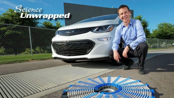 Researcher kneeling beside a white electric Chevrolet Bolt and a coil, with Science Unwrapped title text