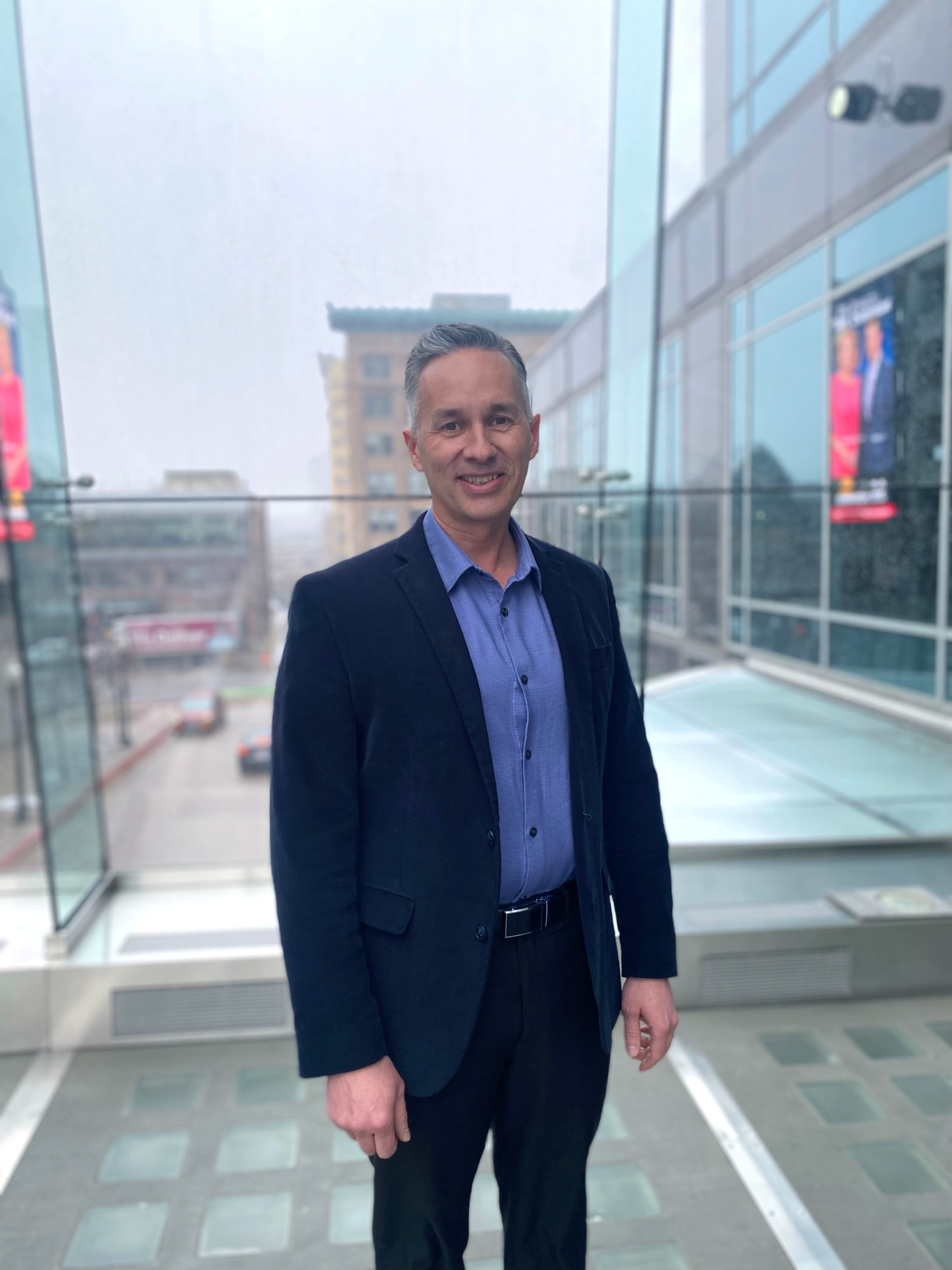 Professional portrait of a man in a dark blazer standing on a glass-walled skywalk overlooking a city