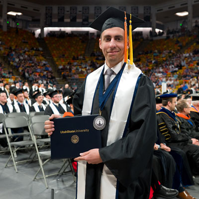 Graduate in cap and gown holding a Utah State University diploma at commencement ceremony