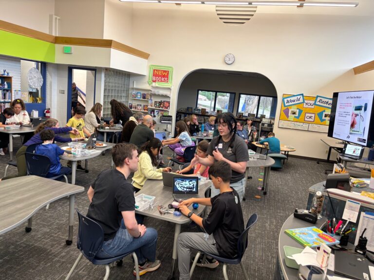 Students and mentors working with laptops and electronics at tables in a school library STEM activity