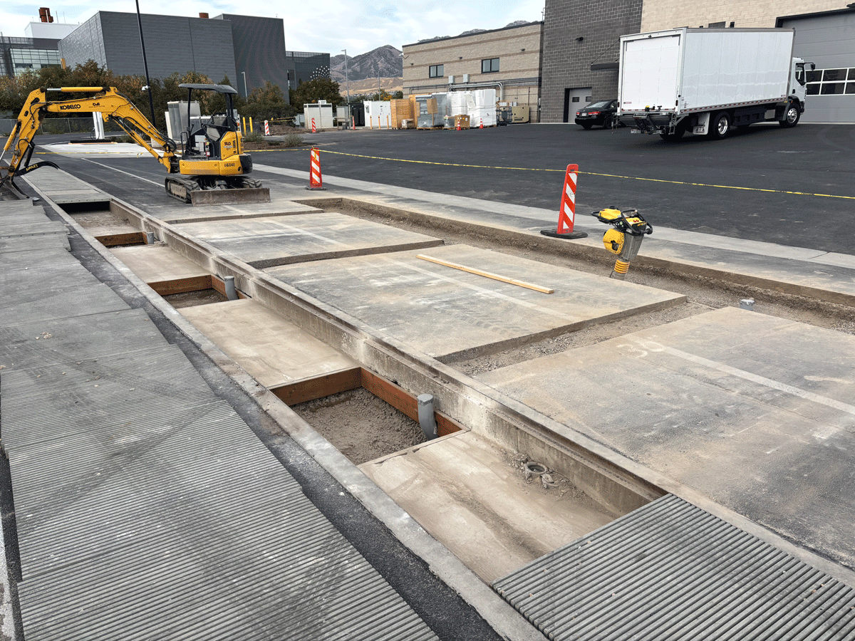 Construction site with mini excavator and exposed concrete trench at a vehicle inductive charging test pad