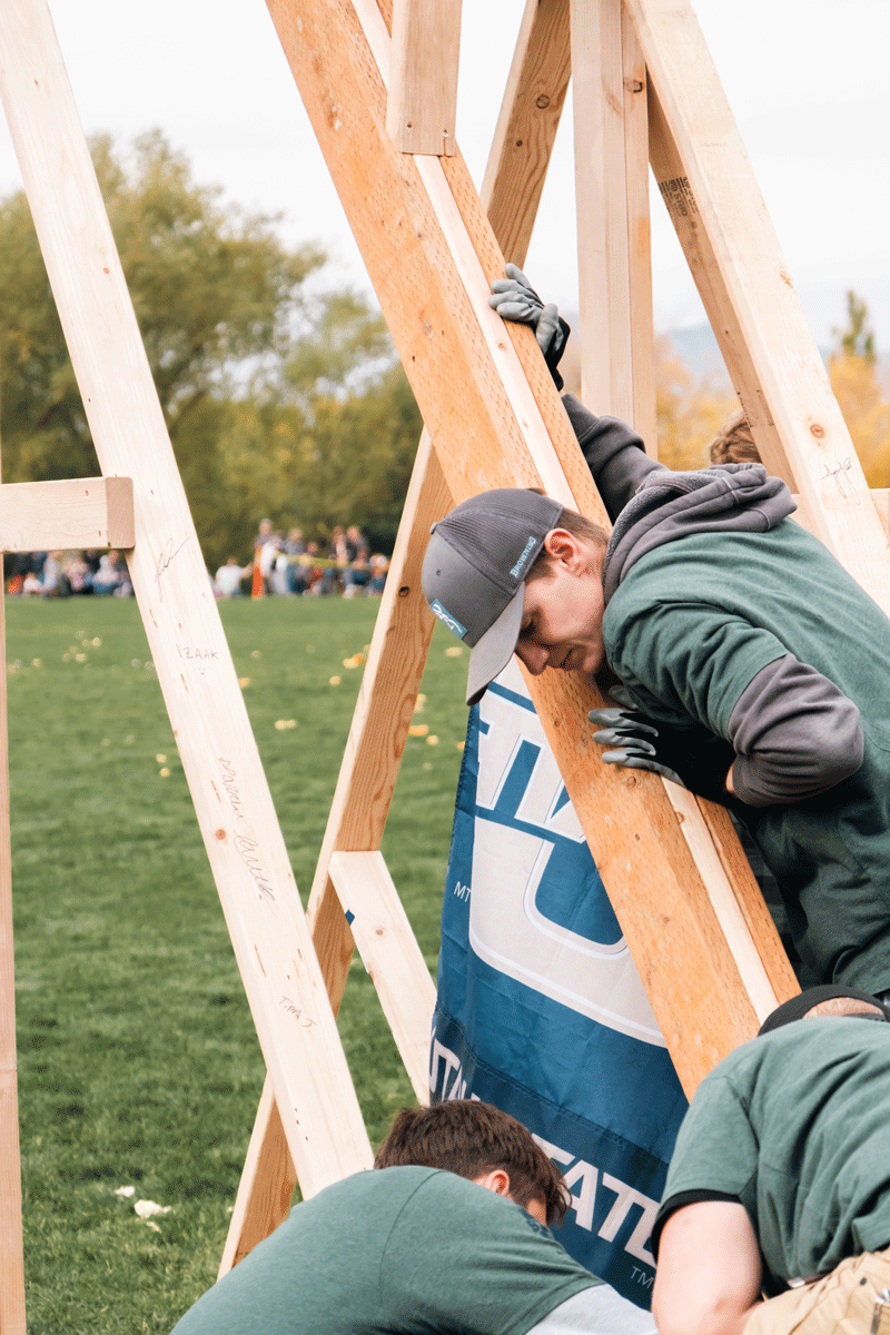 Student in a cap braces a tall wooden trebuchet at an outdoor Utah State University competition