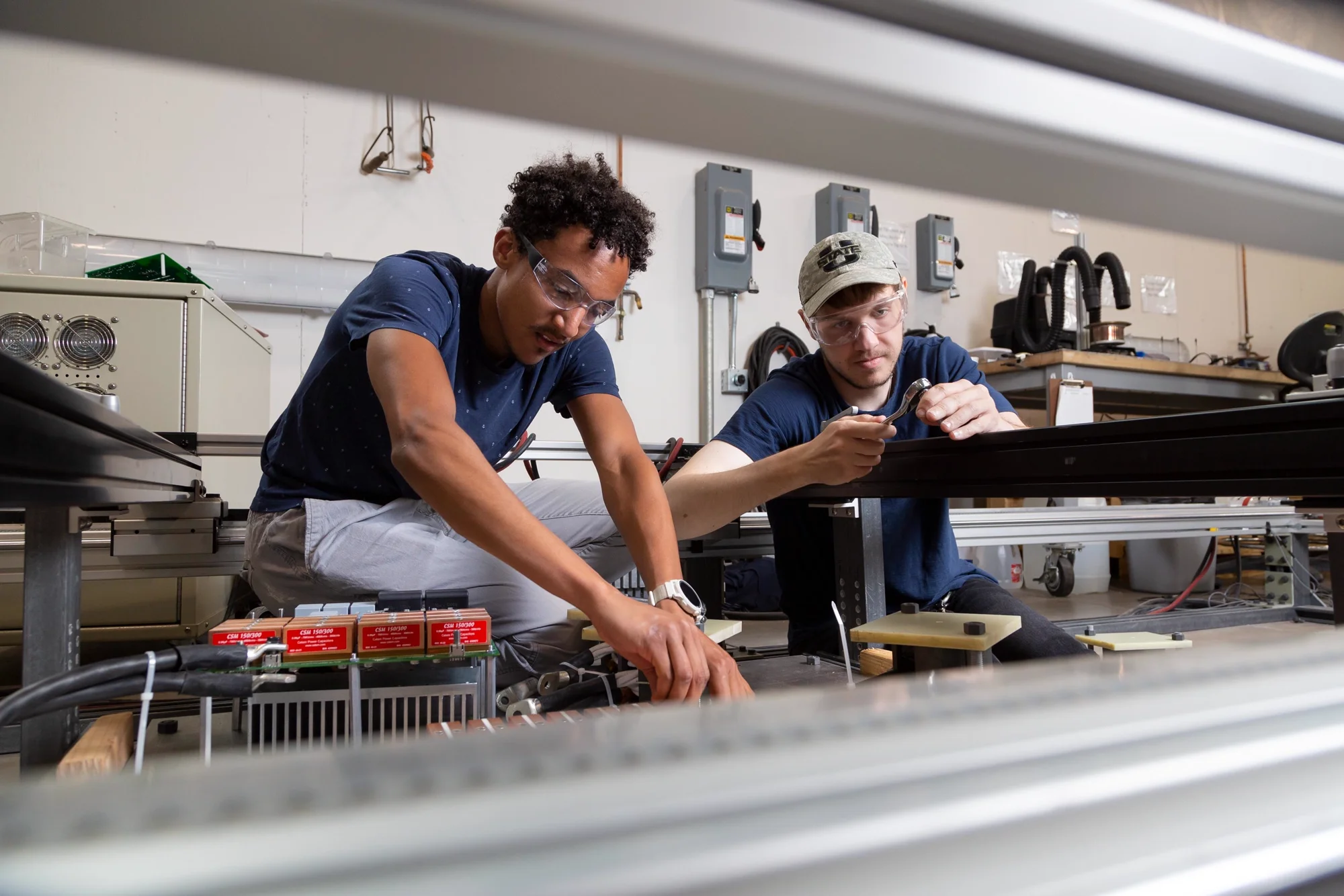 Two students in safety glasses work on electrical equipment along a test track in a lab
