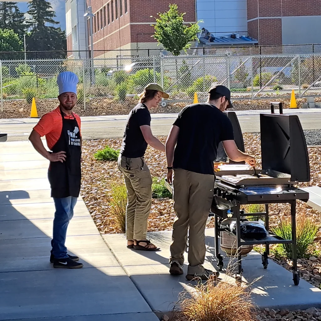 Three men grill food outdoors at a campus cookout, one wearing a tall chef's hat and apron