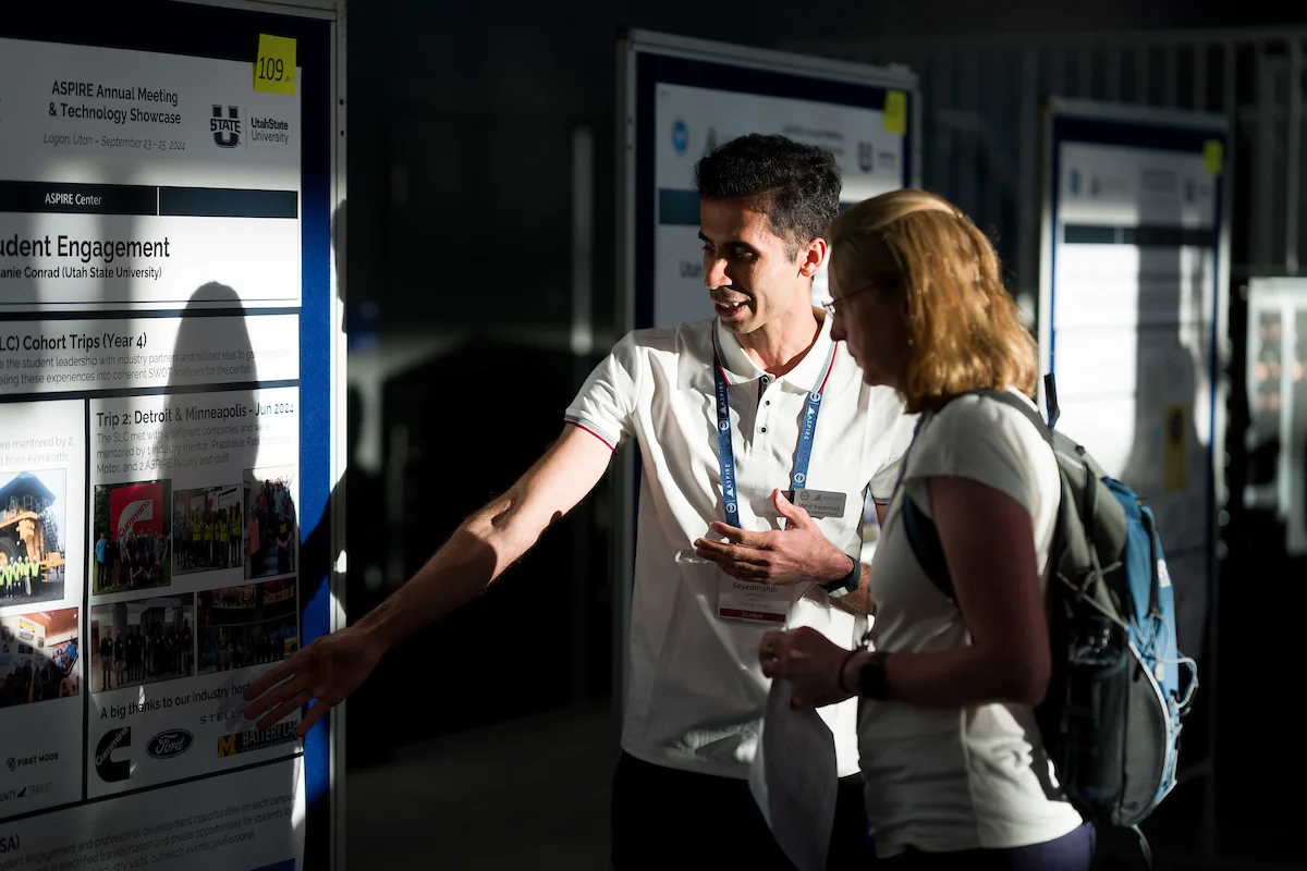 Man in white polo gestures at an ASPIRE Annual Meeting research poster while a woman with a backpack listens