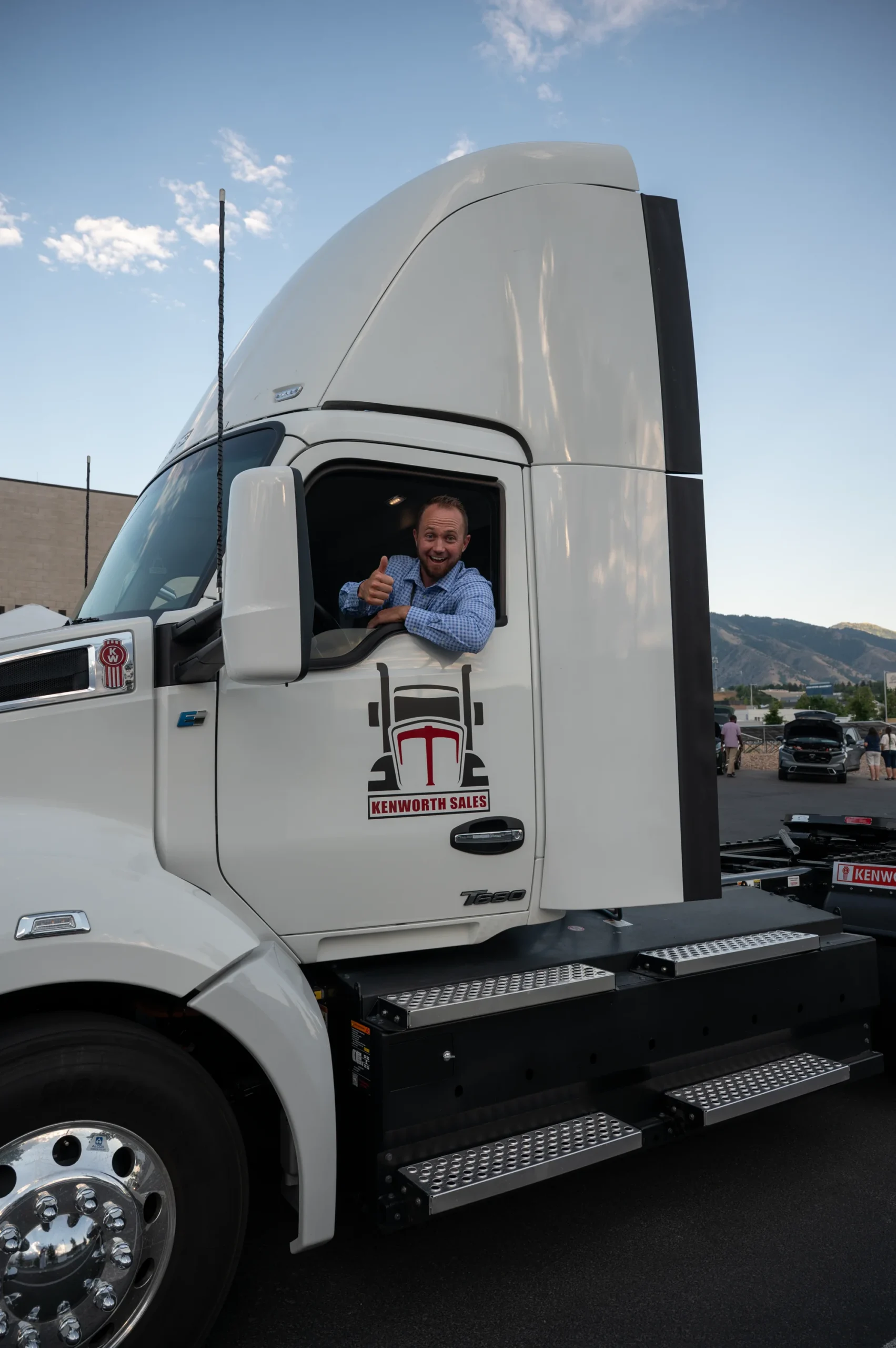 Man gives a thumbs-up from the cab of a white Kenworth semi truck with mountains behind