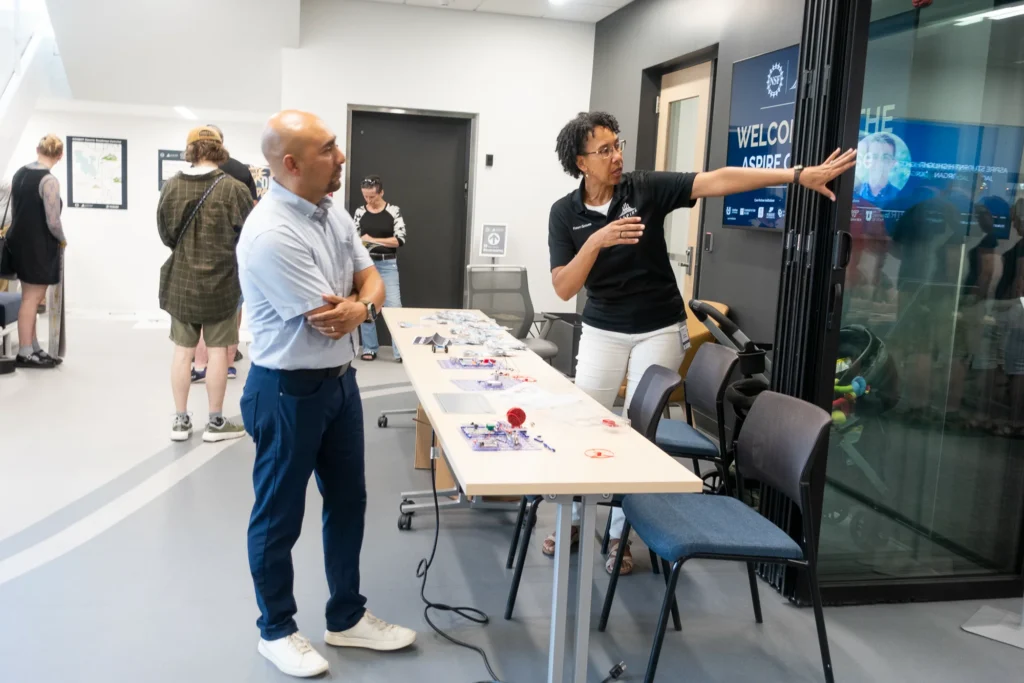 Woman gesturing toward a welcome sign explains the ASPIRE event to a man beside a display table