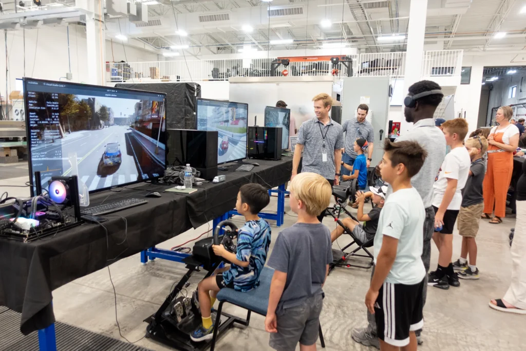 Children using driving simulators with steering wheels at an indoor STEM event while staff supervise.