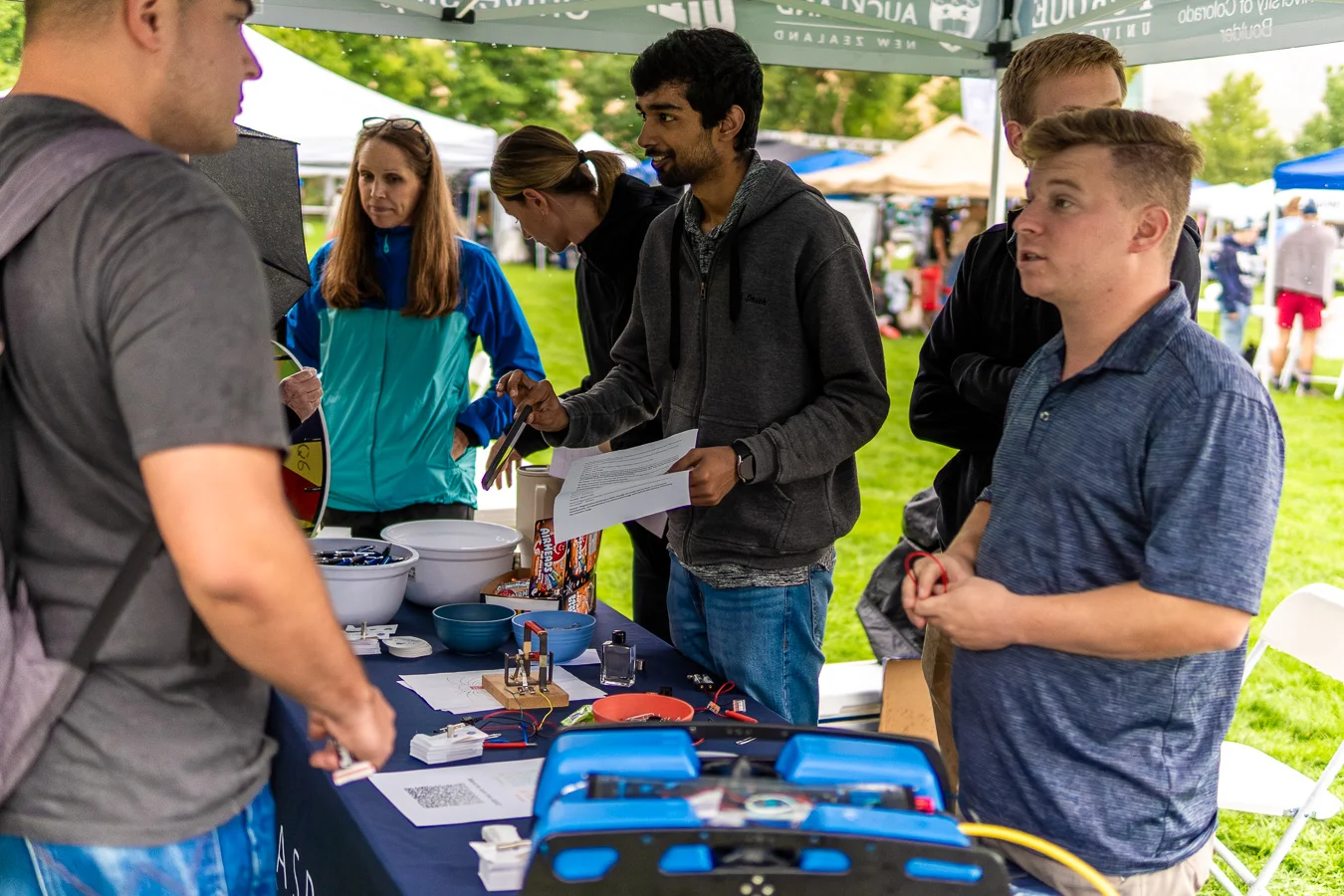 Students demonstrate a project with equipment at an outdoor booth on a grassy campus event
