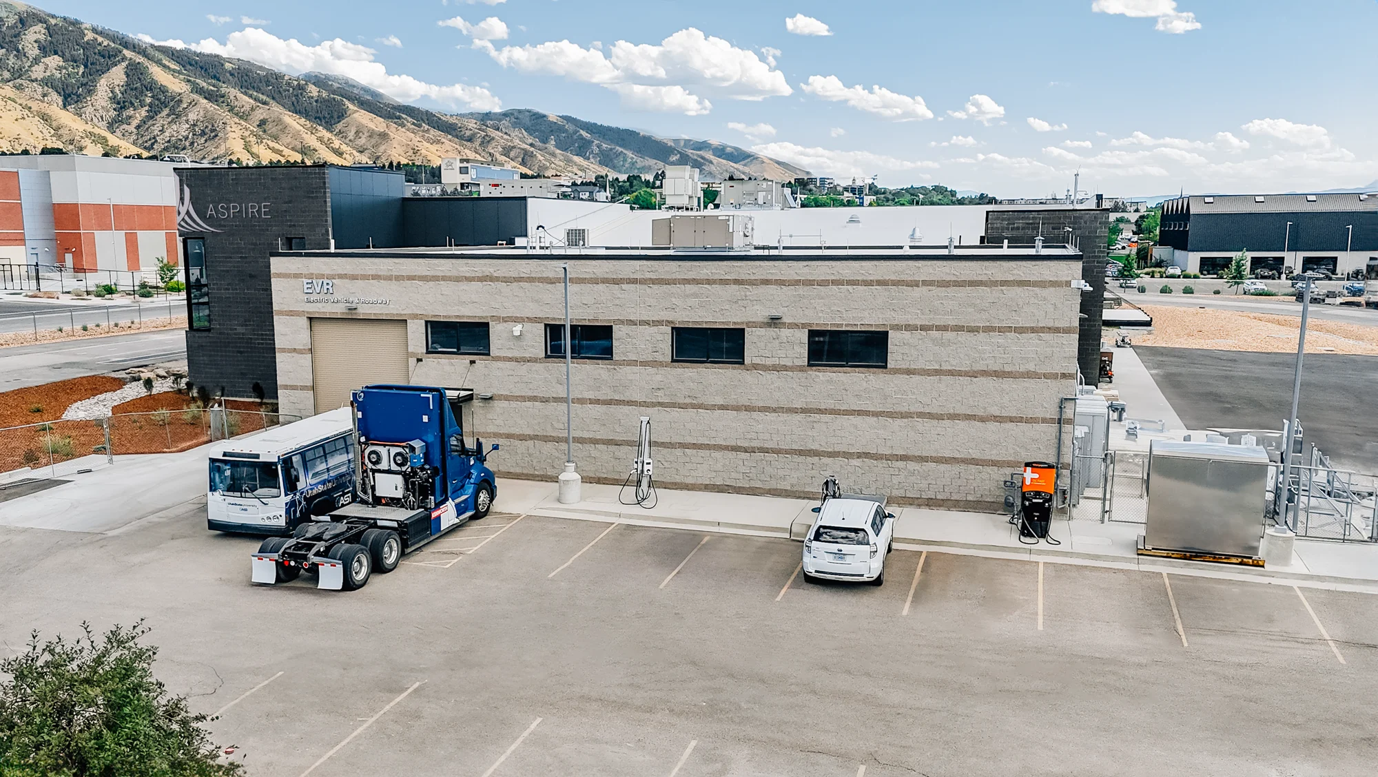 Aerial view of the ASPIRE building with a parked semi-truck and EV chargers, mountains behind.