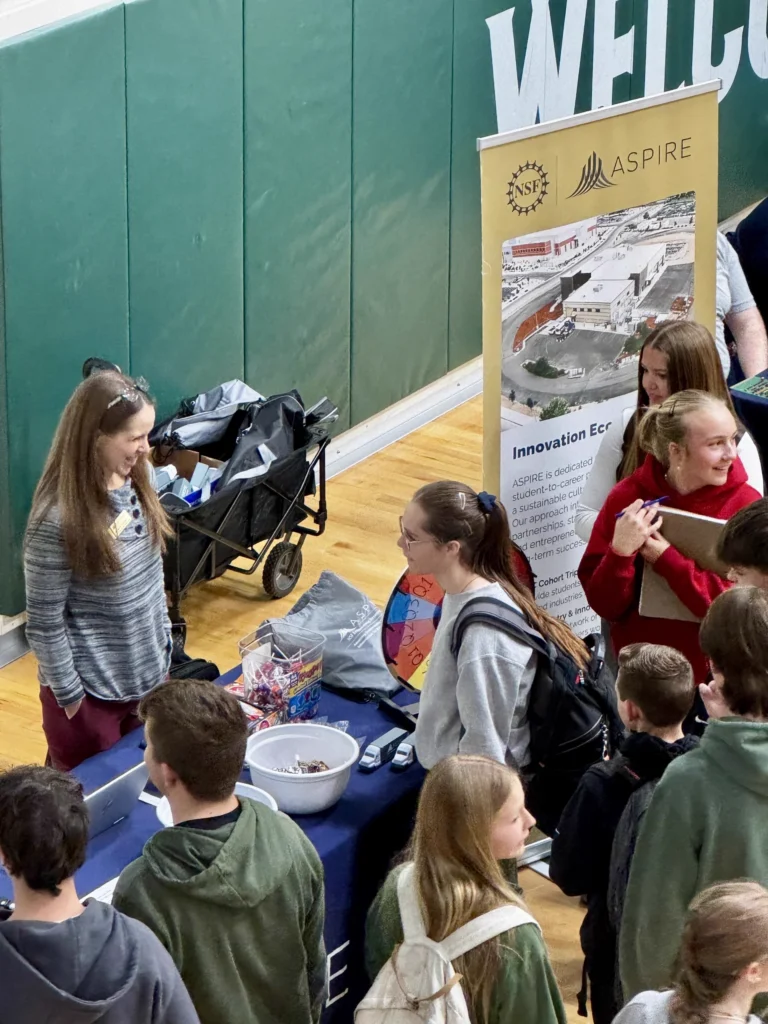Students talking with ASPIRE booth staff at an outreach event in a school gymnasium.