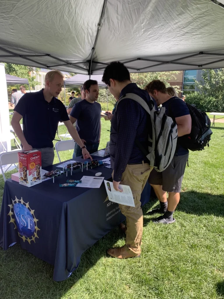 Students examining demonstrations at an outdoor NSF-branded booth tent on a campus lawn.
