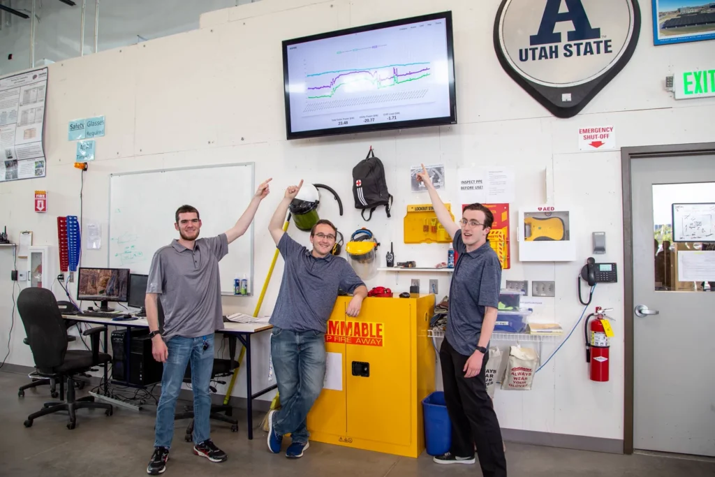 Three young men in a research lab pointing at a wall-mounted monitor showing a data chart.