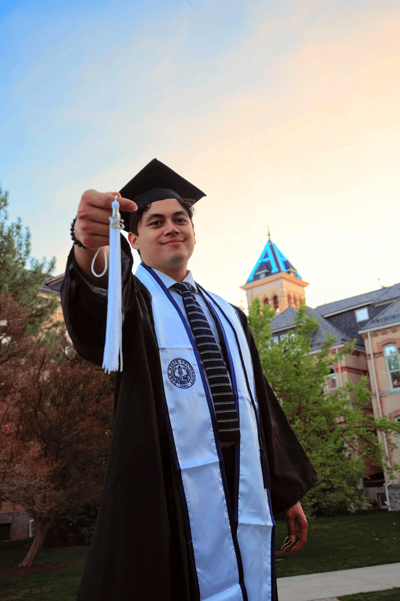 Graduate in cap and gown holding out tassel in front of the Utah State University Old Main tower
