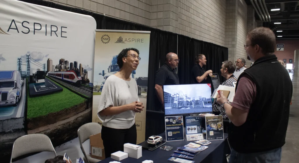 Woman presenting at an ASPIRE exhibition booth with a model train display at a summit.