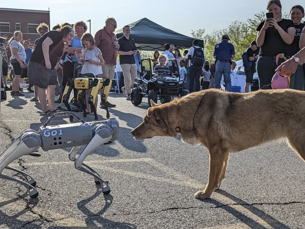 Quadruped robot dog facing a real dog as a crowd watches at an outdoor event.