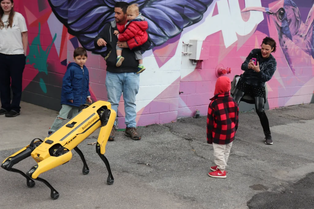 Yellow quadruped robot walks past families viewing a colorful mural at an outdoor event.