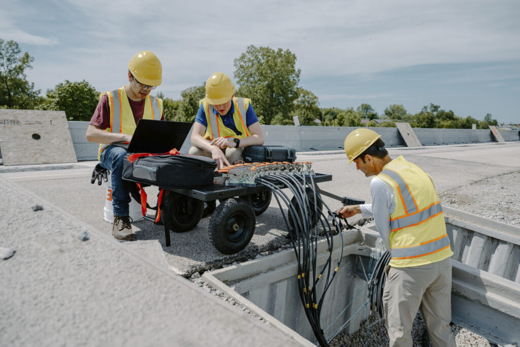 Three workers in hard hats and safety vests testing instrumented sensor cables on a concrete roadway