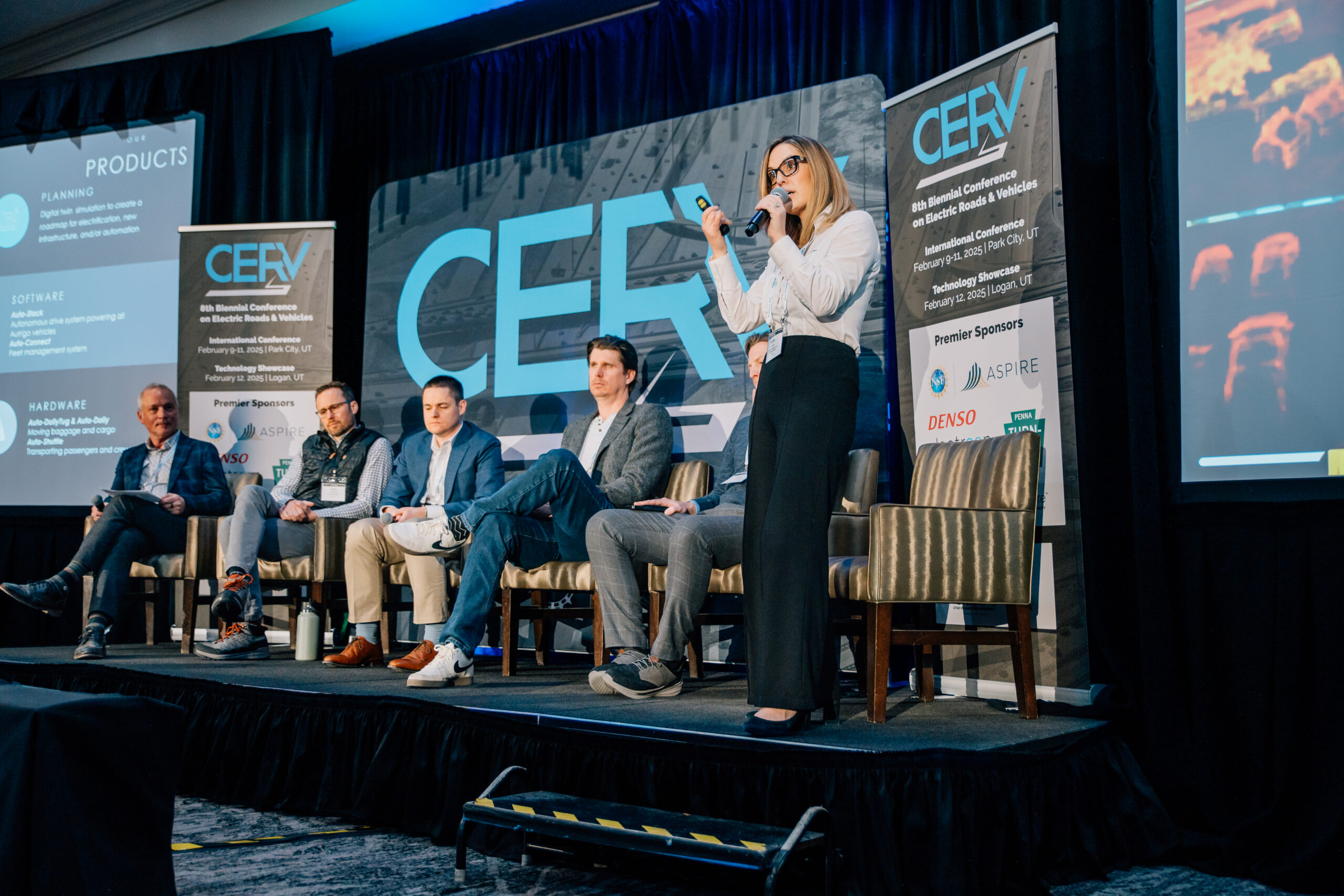 Woman speaking into a microphone on stage beside a seated panel at the CERV electric roads conference