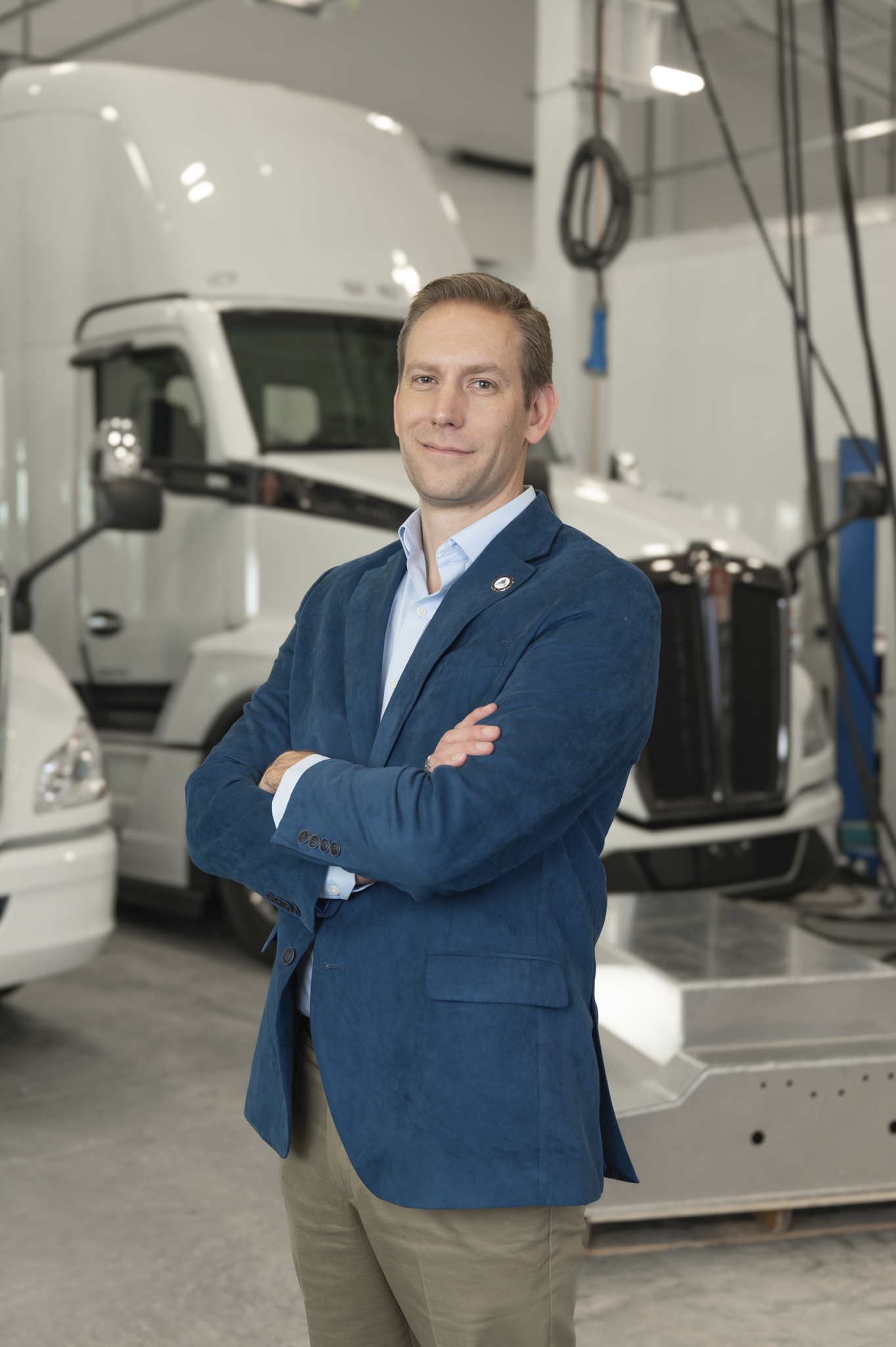 Portrait of Dustin in a blue blazer, arms crossed, in front of white semi-trucks in a lab