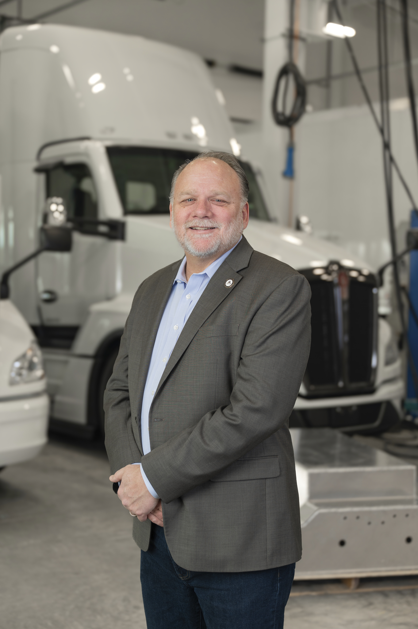 Professional portrait of James, a smiling man in a gray blazer standing before white semi-trucks in a shop